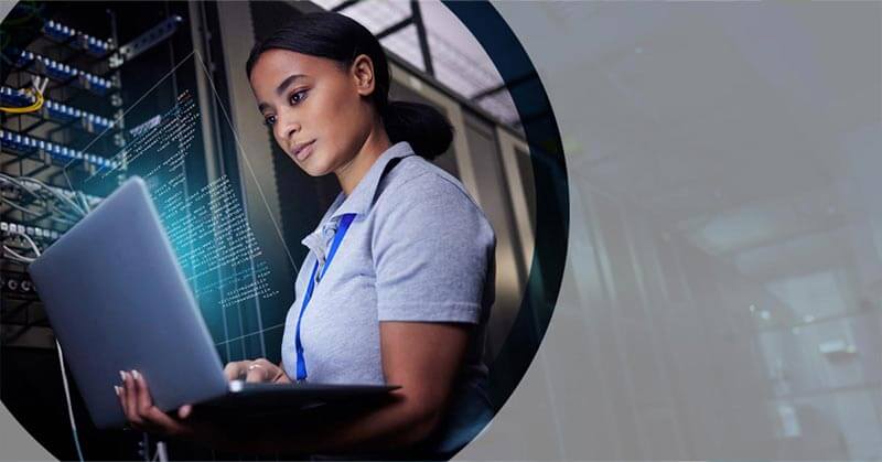 Photo of a woman standing in a server room looking at a laptop, her face is lit from the glow of the screen in the dark room.