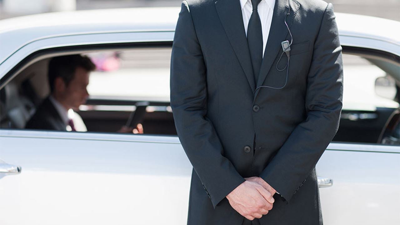 Close-up of a security professional in a black suit and tie standing in front of a white luxury vehicle, wearing an earpiece. An executive sits inside of the vehicle, focused on a tablet device. The scene indicates executive protection, vigilance, and high-level security services.