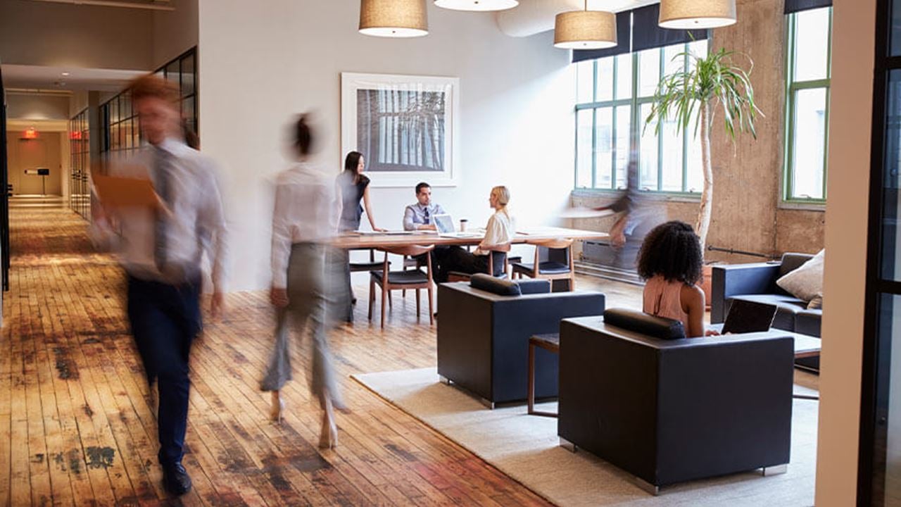 Modern public space with wooden floors, lounge chairs, and a table. Several people are sitting and walking through the area, with pendant lights overhead and large windows letting in natural light. A tall indoor plant is near the windows.