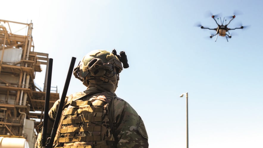 A soldier in camouflage gear and helmet looks up at a quadcopter drone flying in the sky near an industrial structure. The scene suggests military or security use of drone technology.