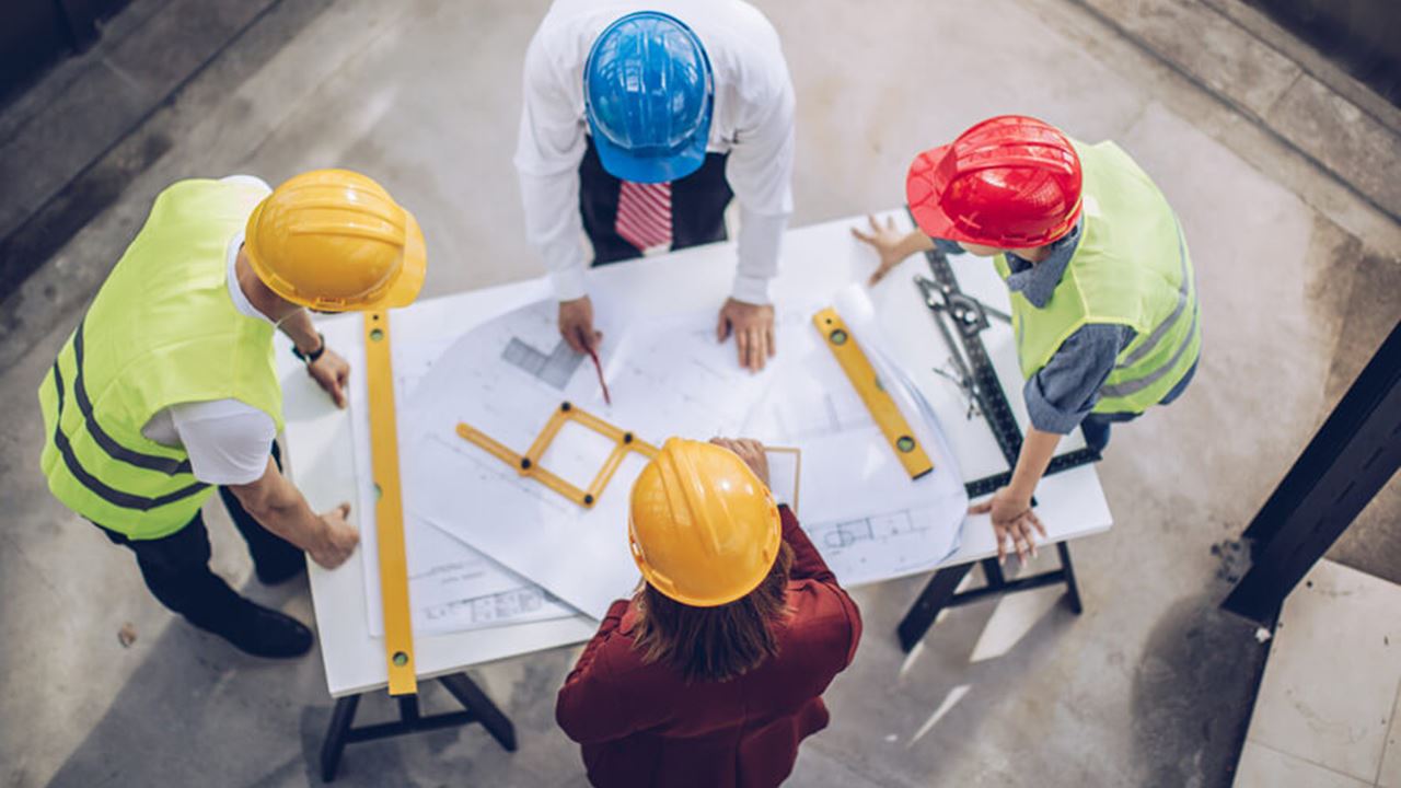 Four construction workers in safety gear review blueprints and tools at a table.