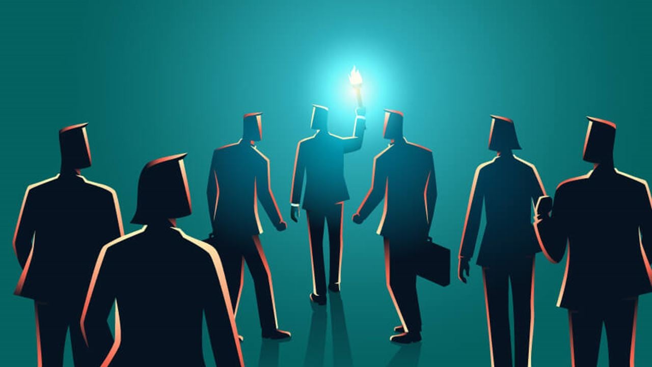 Silhouetted business figures stand in a semi-circle around a central person holding a glowing blue torch, symbolizing leadership, guidance, and enlightenment amid dark or frightening times.