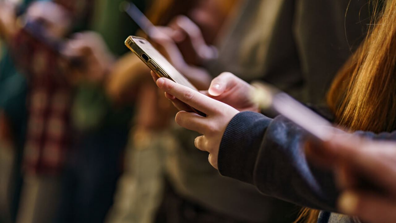 Close-up photo of several teenagers holding and using smartphones, with one phone in sharp focus and others blurred in the background.
