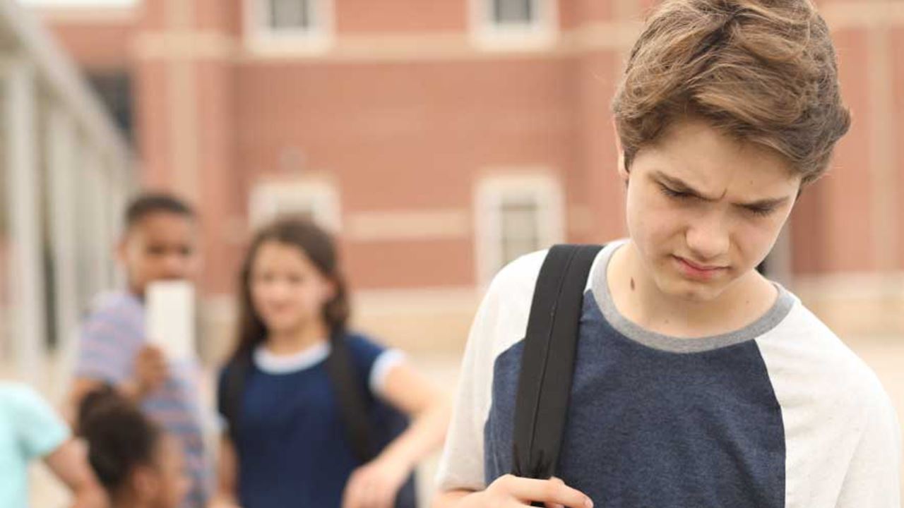 Middle school student with a backpack looking downward and sad or concerned in the foreground, with several other students talking about the boy outside a school building.