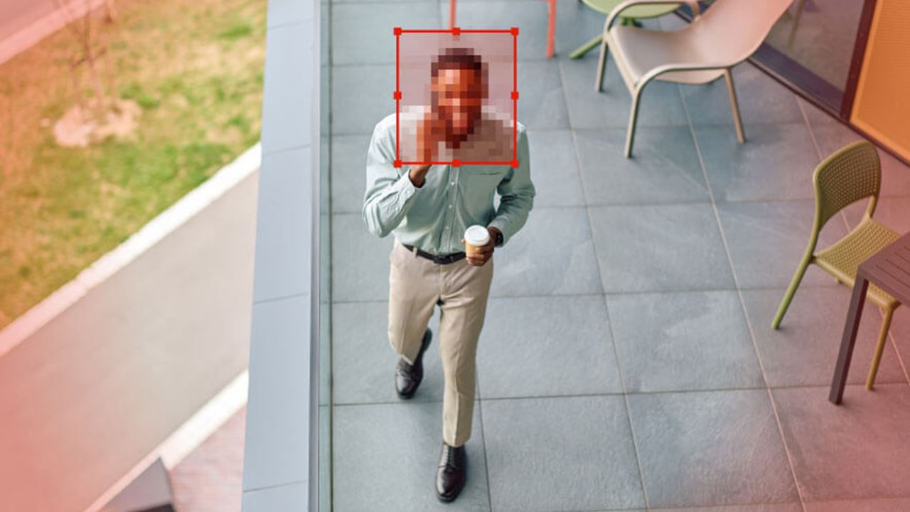 Overhead view of a person walking on a patio holding a coffee, with the face pixelated and outlined by a red detection box, suggesting automated facial recognition or privacy masking.