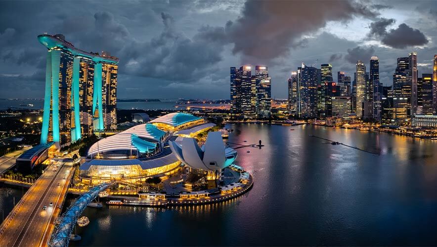 Evening view of Singapore’s Marina Bay waterfront, showing the illuminated Marina Bay Sands hotel with three towers and a sky park, the lotus-shaped ArtScience Museum, and a city skyline of high-rise buildings reflected in calm water. The sky is dramatic with dark clouds.