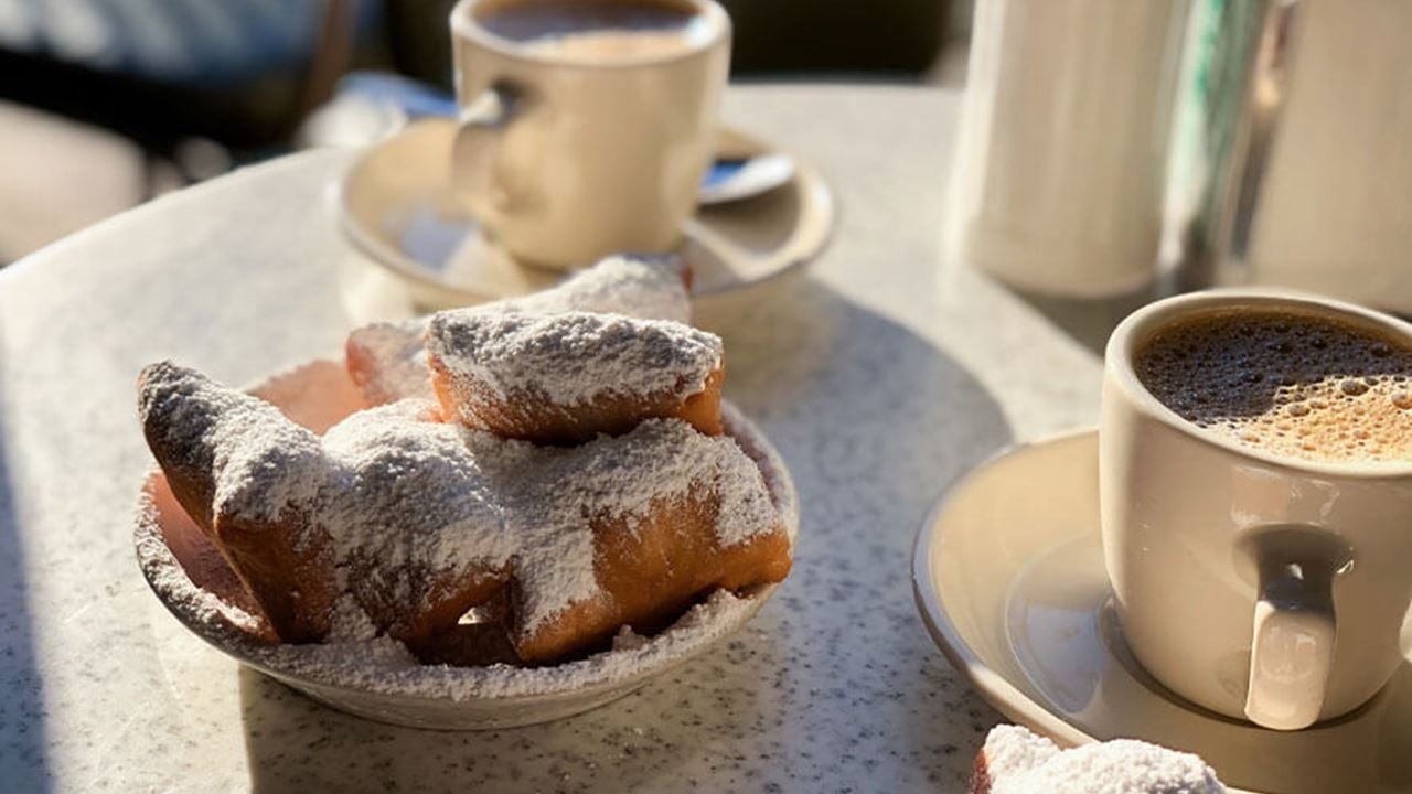 A plate of powdered sugar-covered beignets on a table, accompanied by two cups of coffee. A sugar dispenser and napkin holder are visible in the background.