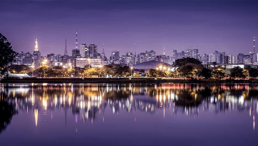 Nighttime view of São Paulo’s skyline with illuminated buildings and towers reflecting on a calm body of water. The sky has a purple hue, and trees line the waterfront, creating a peaceful urban scene.