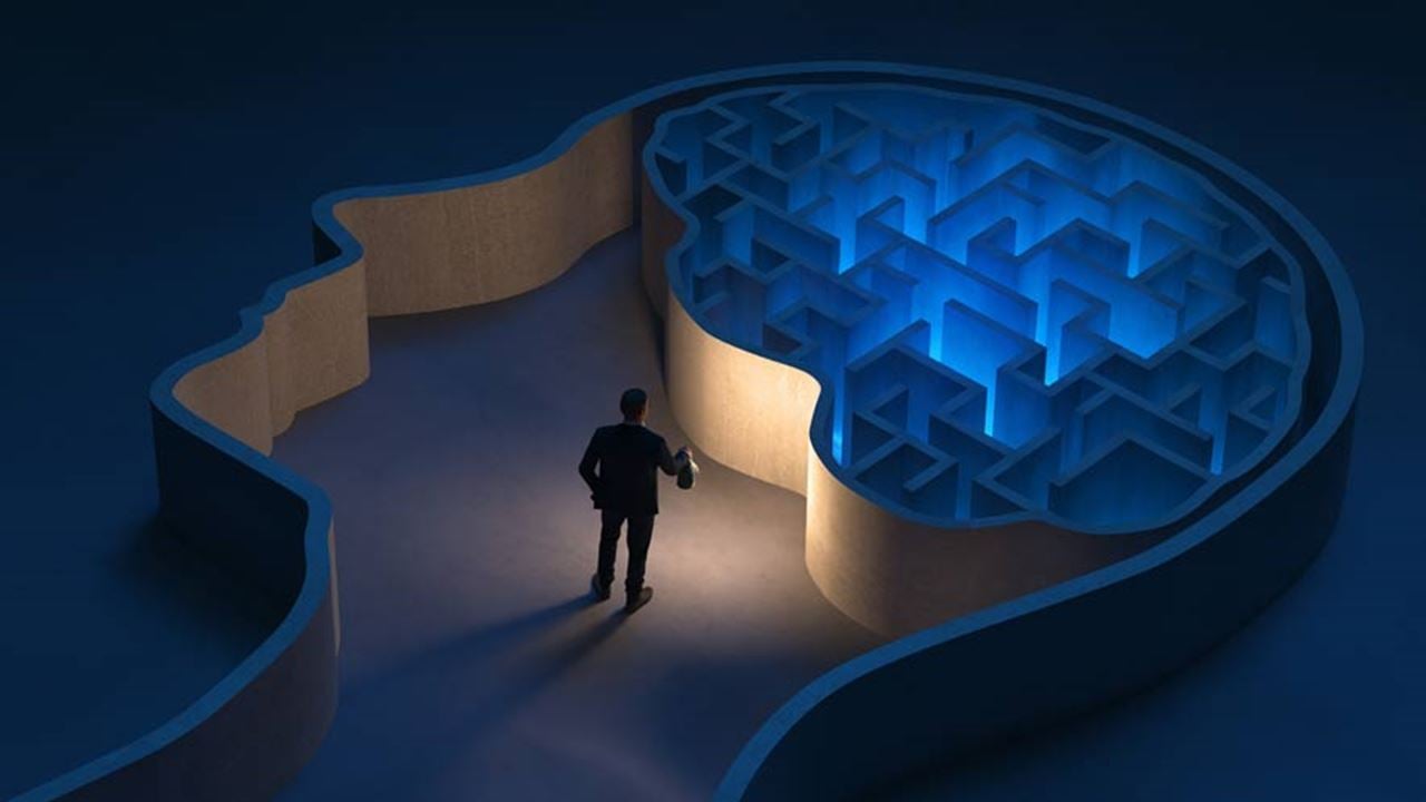 Photo illustration of a male security manager wearing a business suit holding a lit lantern standing inside a maze in the shape of a human head. The man is attempting to make a decision about which direction to take.