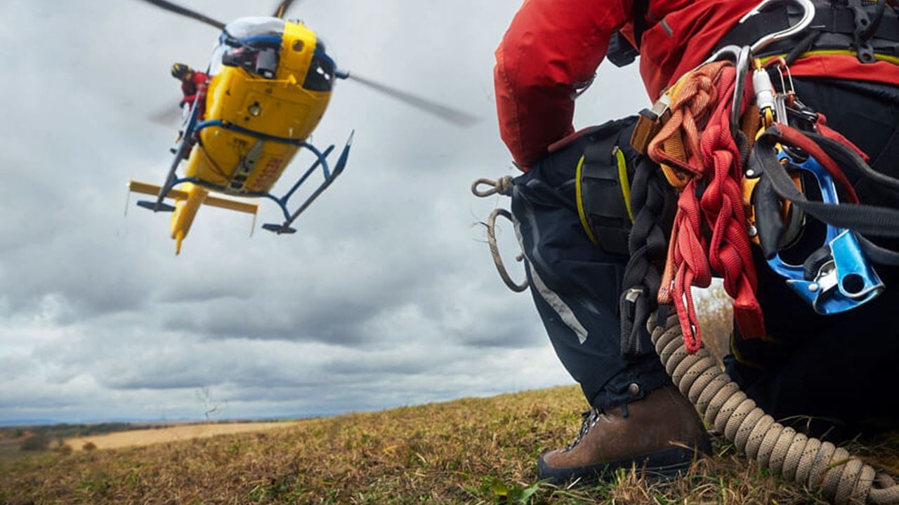 Close-up of safety harness worn by paramedic from emergency service, with a landing helicopter in the background on a hill under a cloudy sky.